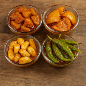 Four small glass bowls filled with traditional Indian pickles including mango, lemon, garlic, and green chilli, arranged on a rustic wooden surface showcasing rich colors and textures for export and culinary use.
